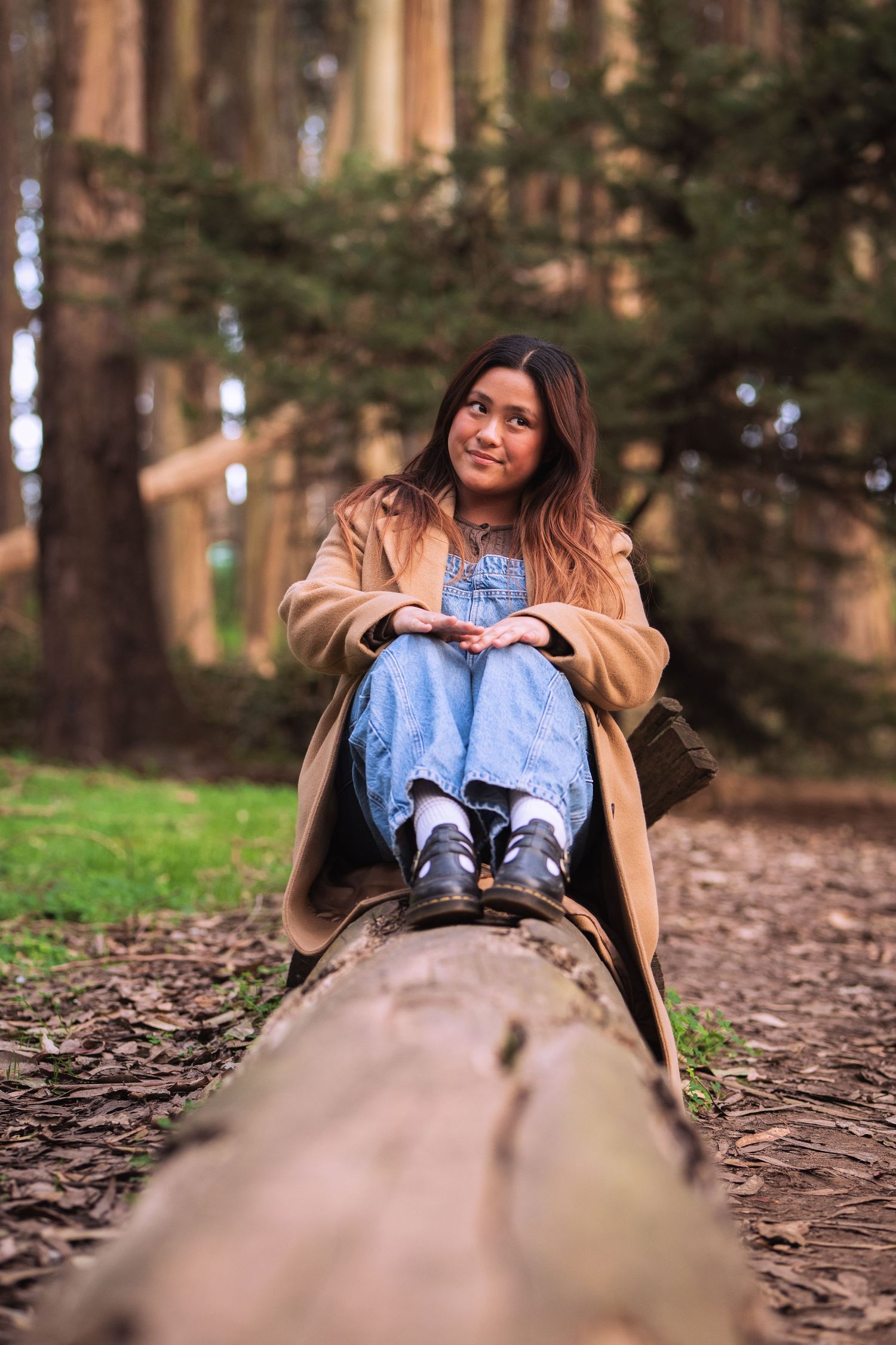 Young woman in beige coat and jeans sitting on a fallen log in a lush green forest, smiling at camera.