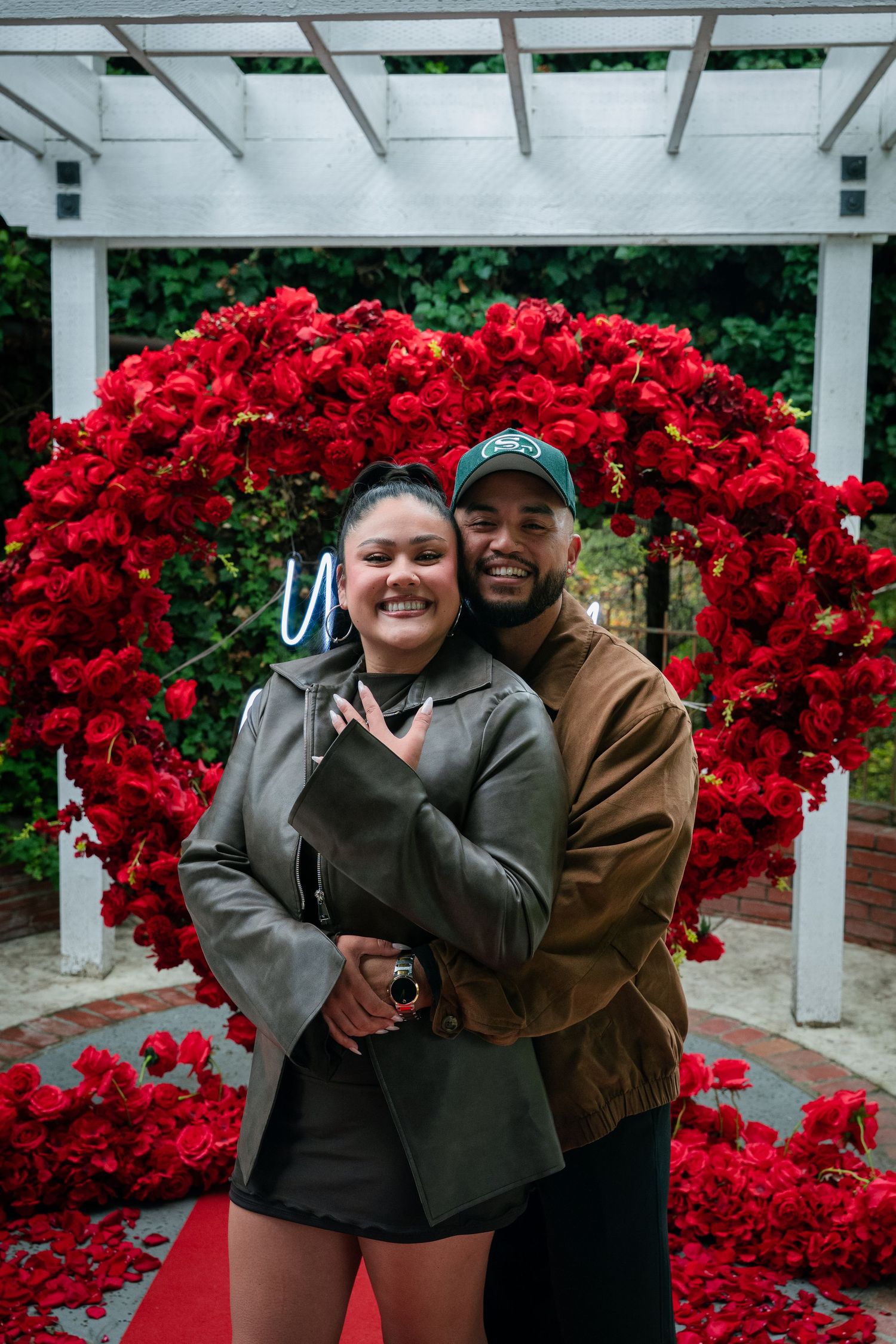 A couple embraces and smiles while standing near vibrant red azalea bushes in a garden setting.