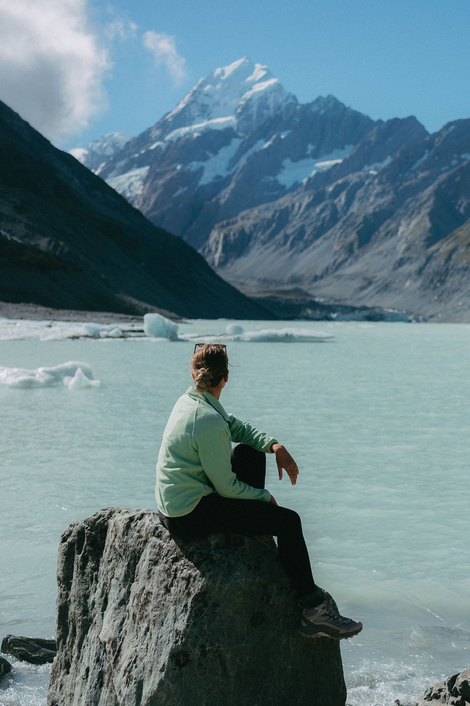 A person sits on a rocky outcrop overlooking the turquoise waters of a glacial lake with snow-capped mountains in the distance.