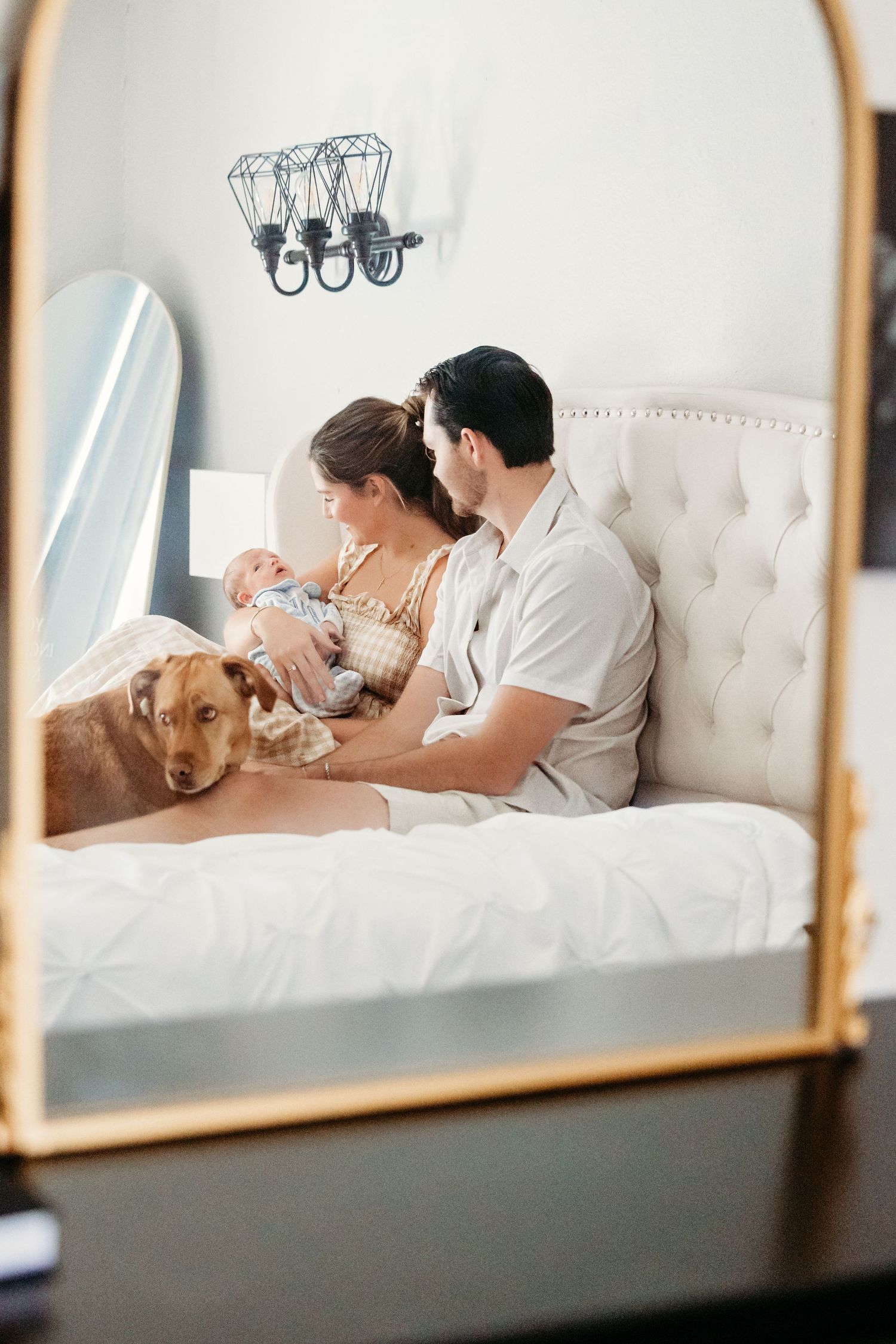 A couple relaxes on a white tufted bed, cradling their newborn with their dog snuggling them in Keller, TX.