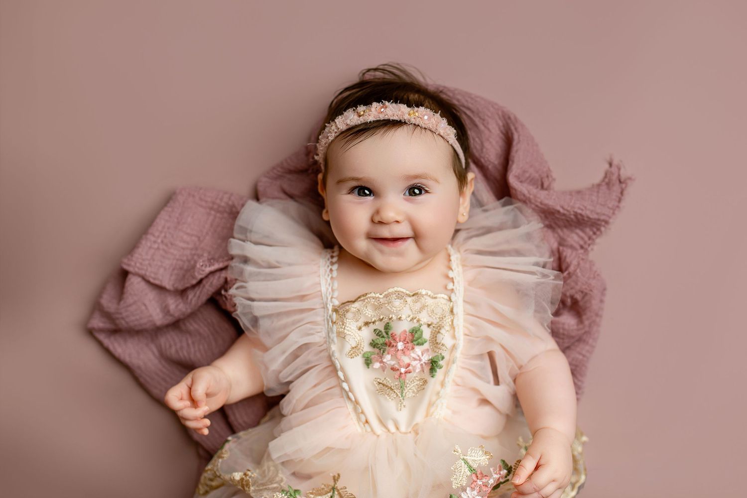 Baby girl in pink embroidered tulle dress with floral headband smiling on mauve backdrop during milestone session in Summerville SC