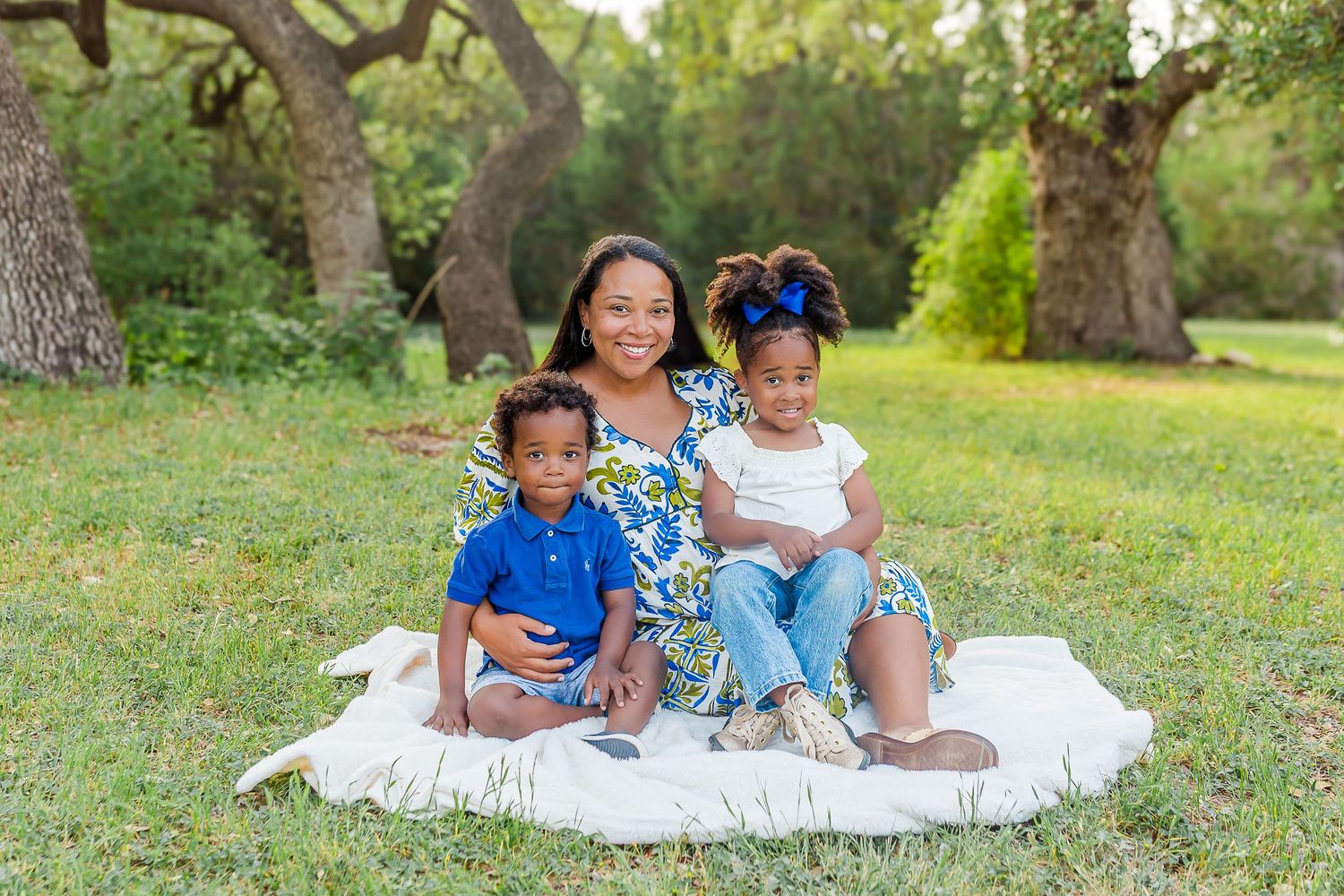 A family enjoys a relaxing picnic together on a white blanket in a lush green park setting.