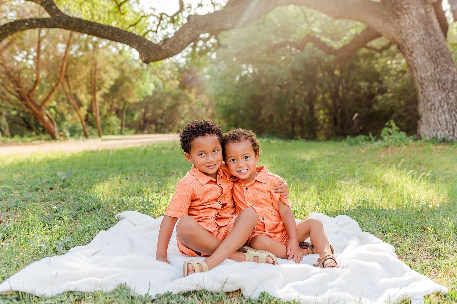 Two young children in matching orange shirts sit together on a white blanket under a tree in a sunlit park.