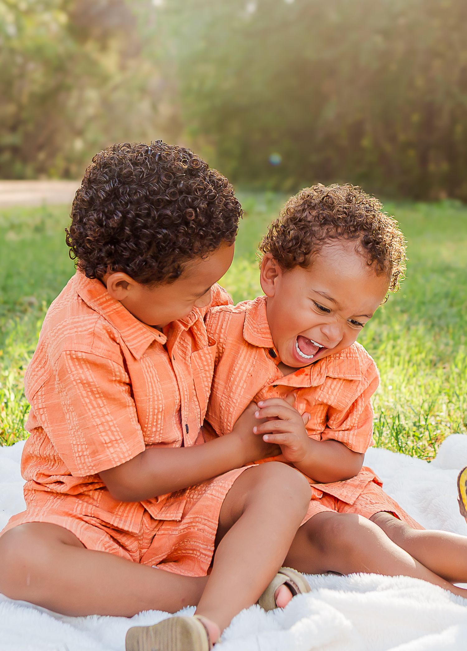 Two children in matching orange shirts laugh and play together on a white blanket in a grassy field at sunset.