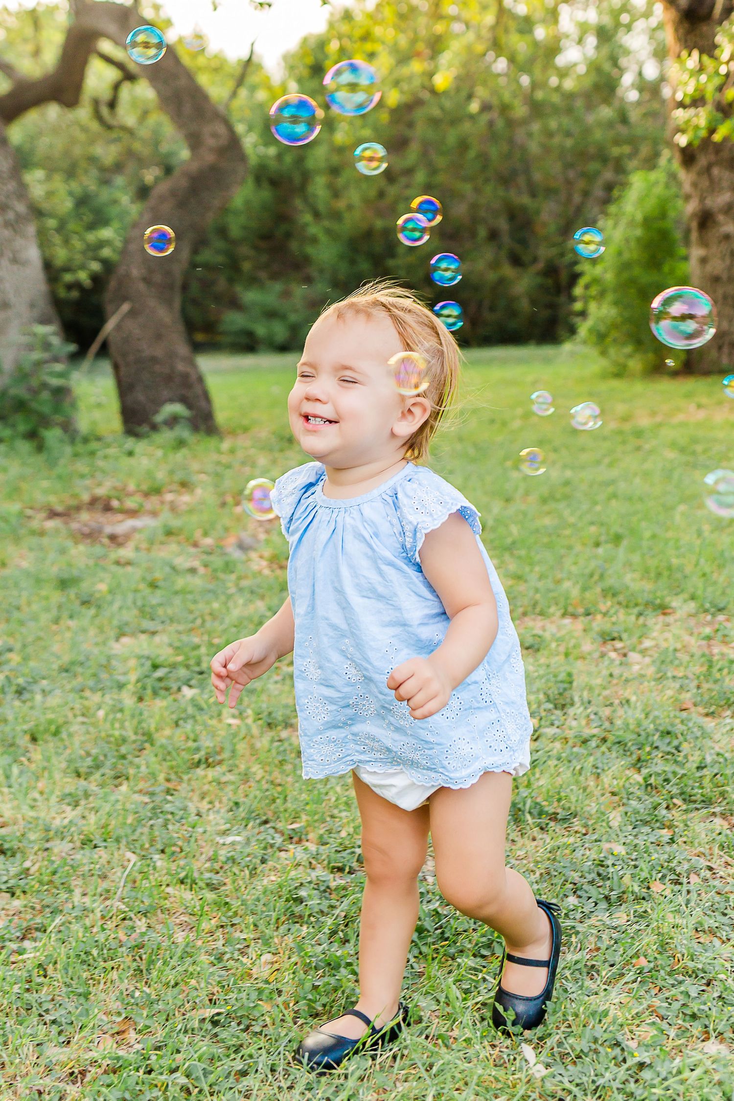 A toddler in a blue dress chases soap bubbles while playing in a grassy backyard on a sunny day.