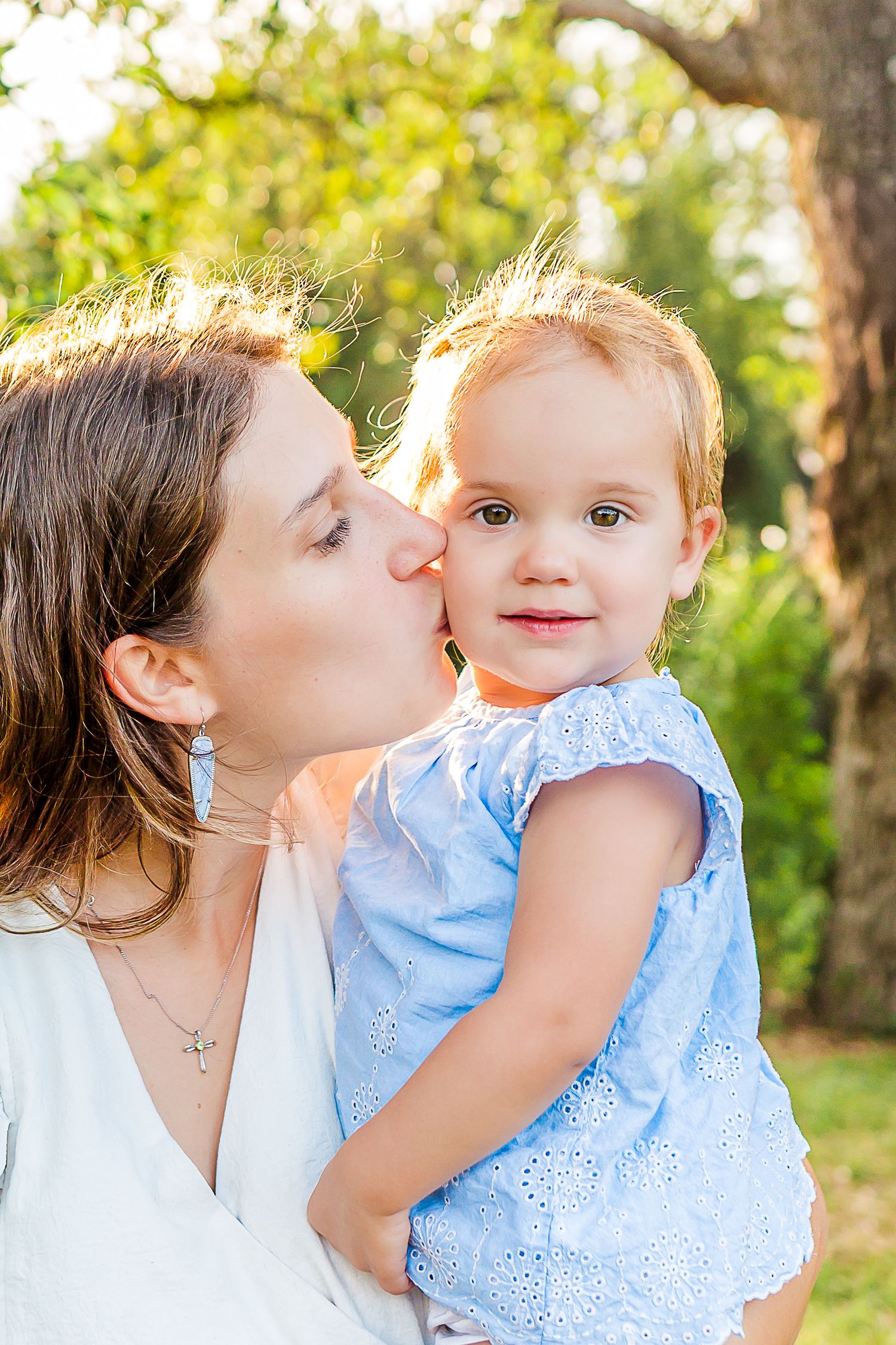A tender moment between parent and child outdoors in a sunlit garden setting, wearing white and light blue outfits.