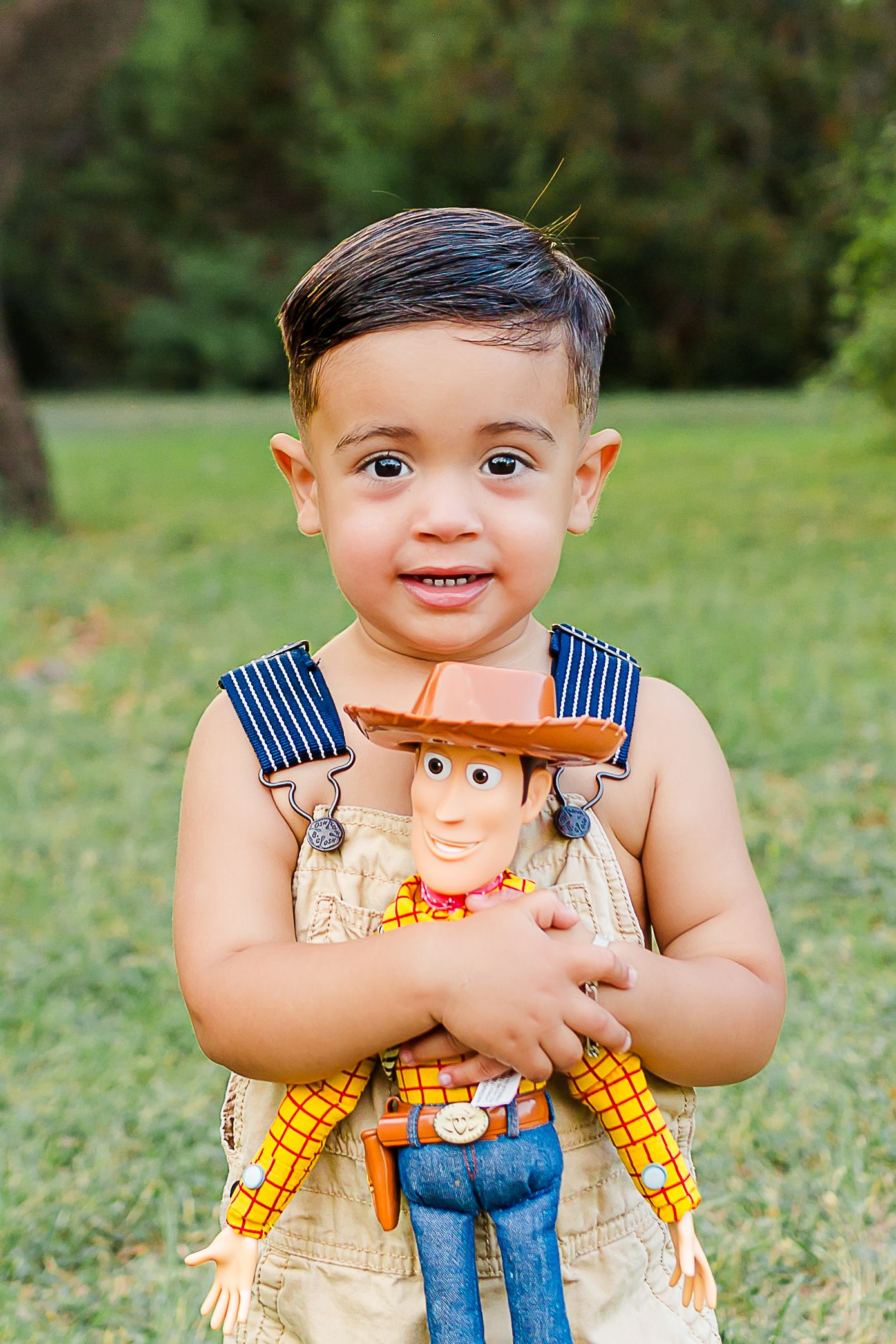 A child in overalls holds a Woody doll from Toy Story while standing in a grassy outdoor setting.