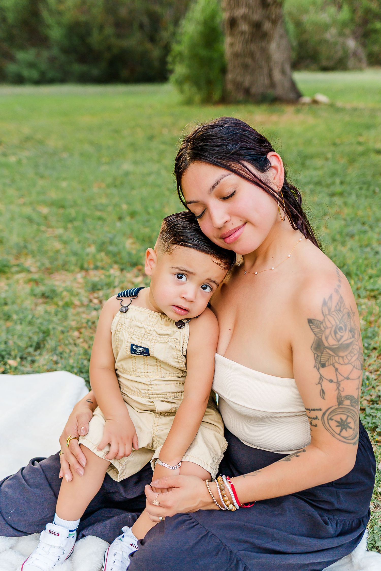 A tender outdoor portrait showing an intimate moment between parent and child sitting together on a blanket in a park.