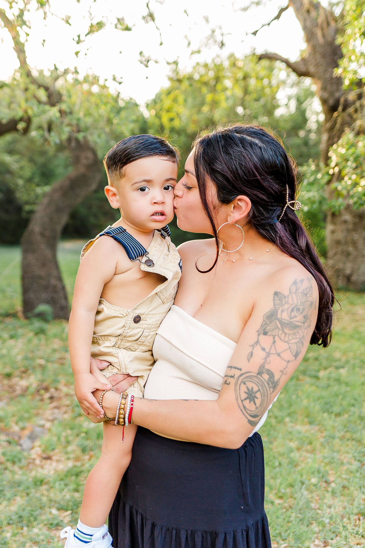 A mother in a black skirt and white top holds her young child wearing tan clothing in a sunny outdoor setting.