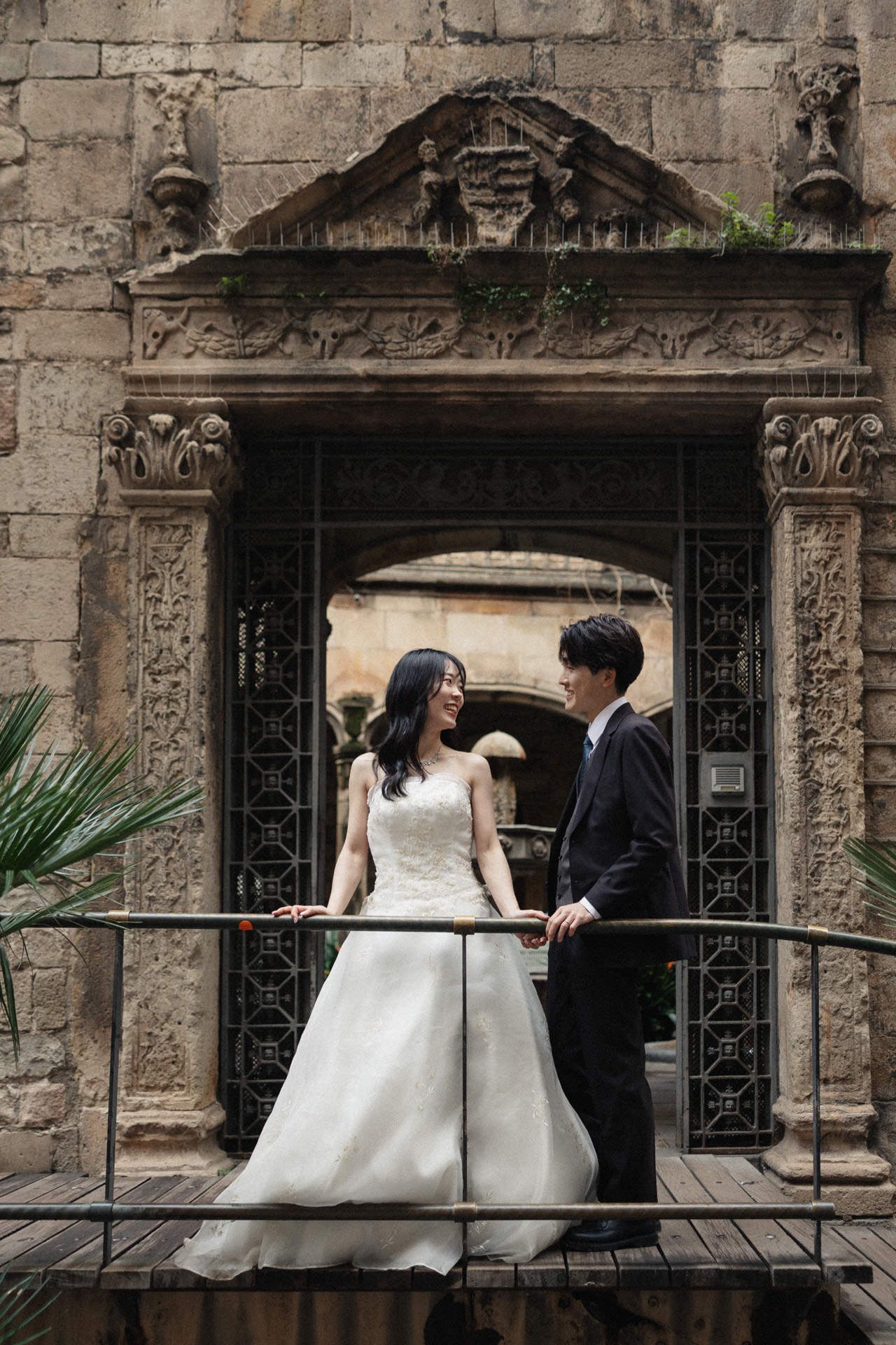 A couple in wedding attire shares an intimate moment against a rustic stone wall with ornate architectural details.