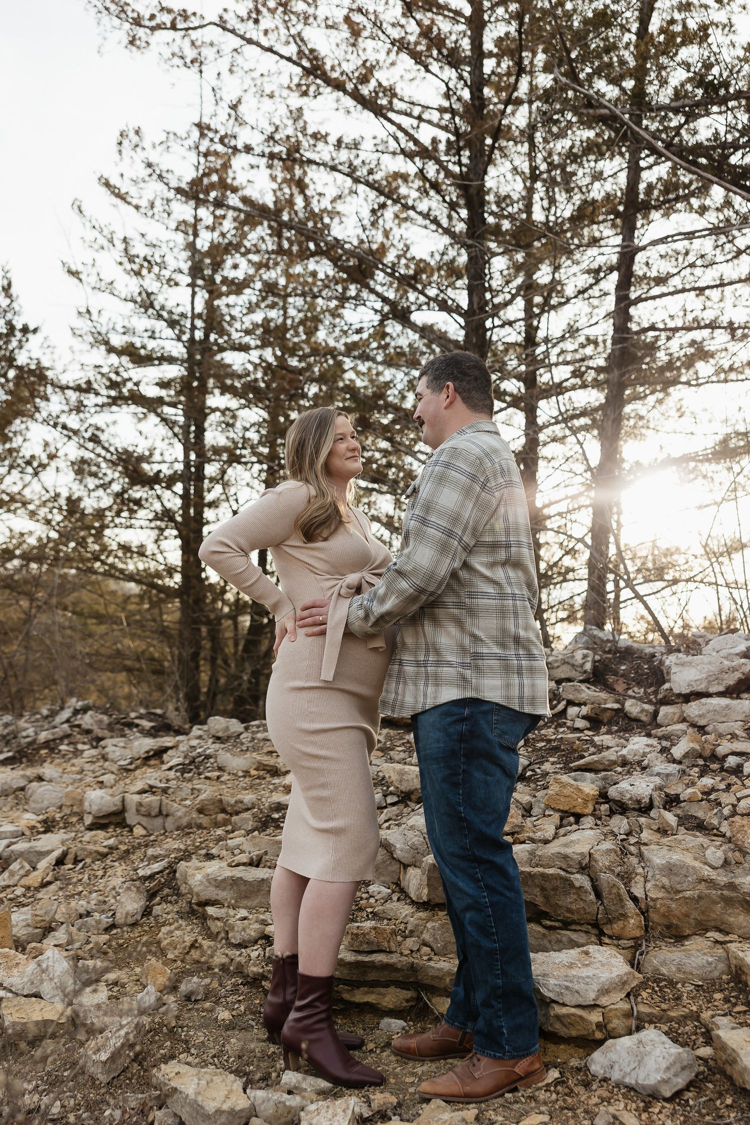 pregnant mother to be stands in front of pine tress dad holds baby bump during photo session