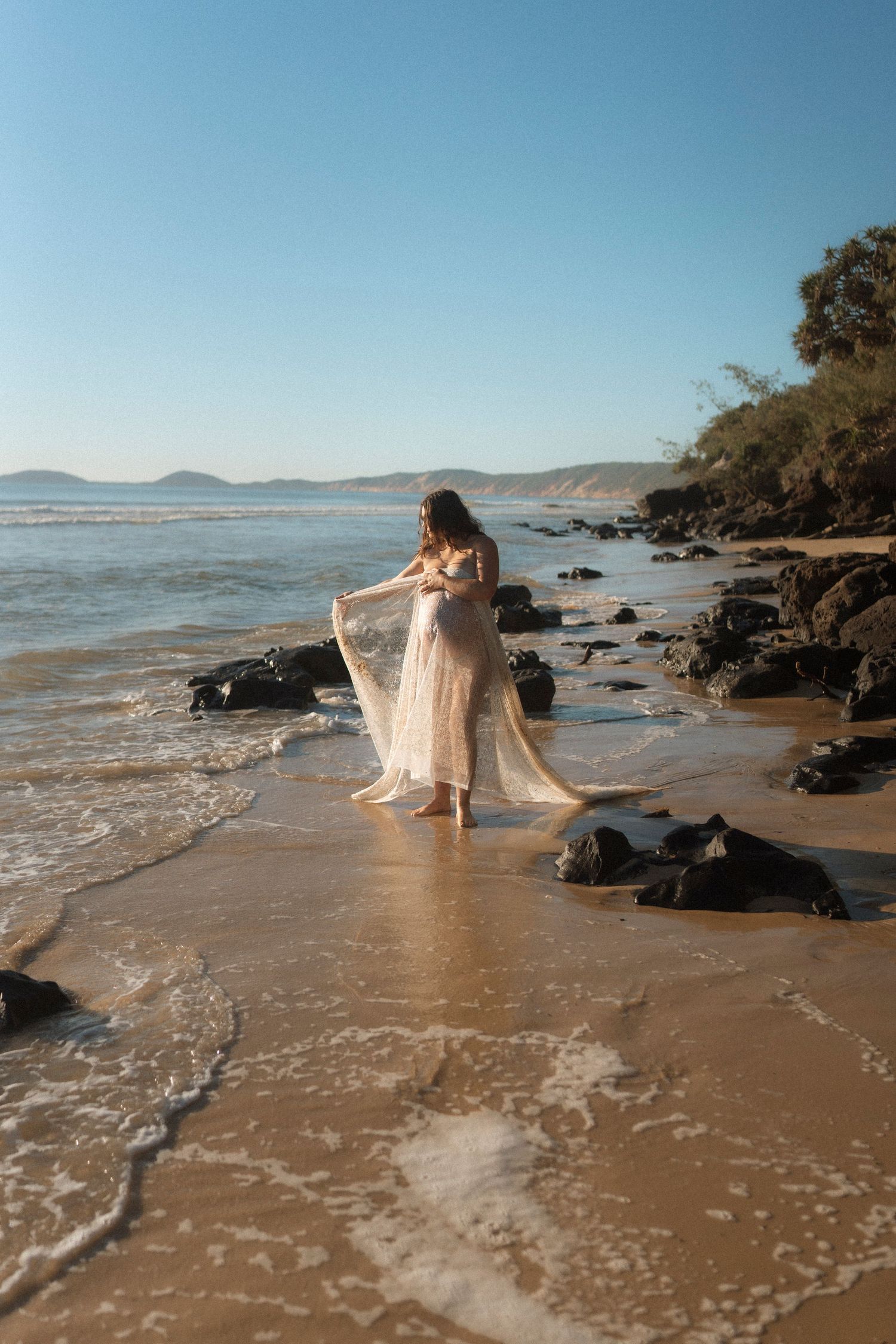 A flowing white dress catches the ocean breeze as waves lap against a rocky beach shoreline at sunset.