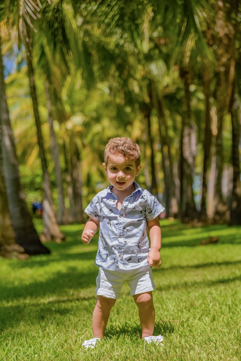 A young child in a patterned shirt and shorts plays and smiles while crouching on green grass in a sunny park.