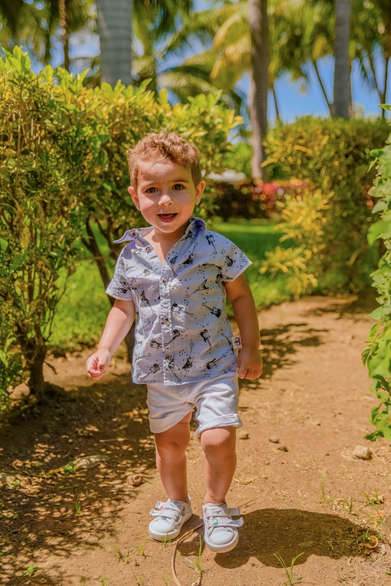 Small child in a patterned shirt and shorts walks along a garden path surrounded by tropical plants and palm trees.