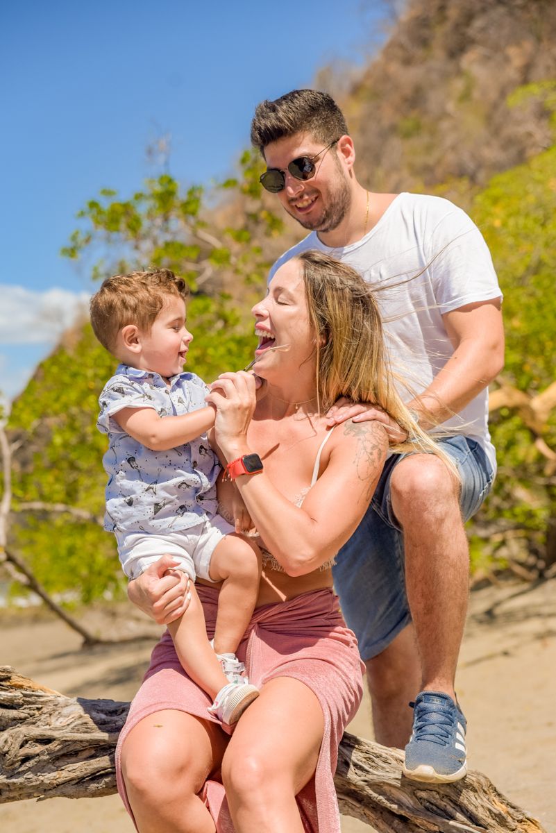 A family shares playful moments together on a sunny beach with rocky cliffs in the background.