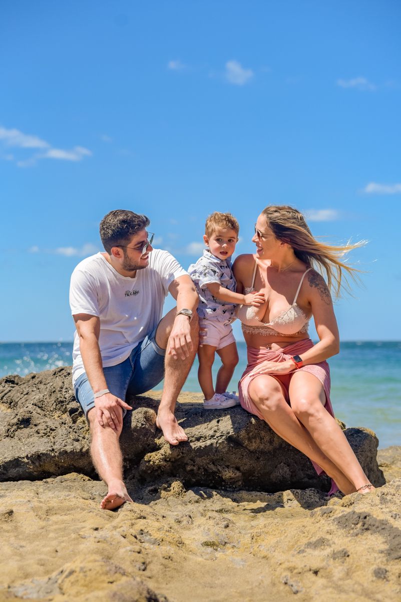 Family sitting together on a rocky beach formation with blue ocean and sky in the background.
