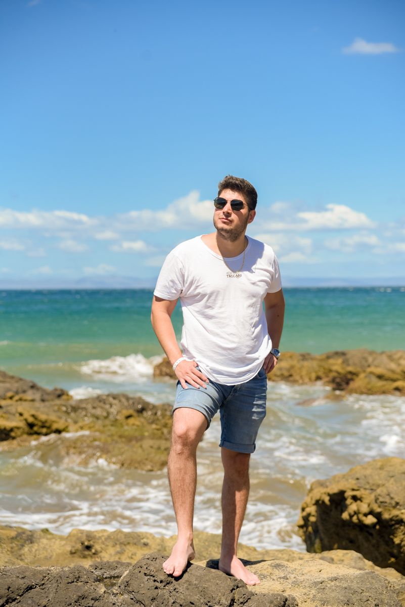Person in white t-shirt and denim shorts enjoying a sunny day at a rocky beach with turquoise ocean waters.
