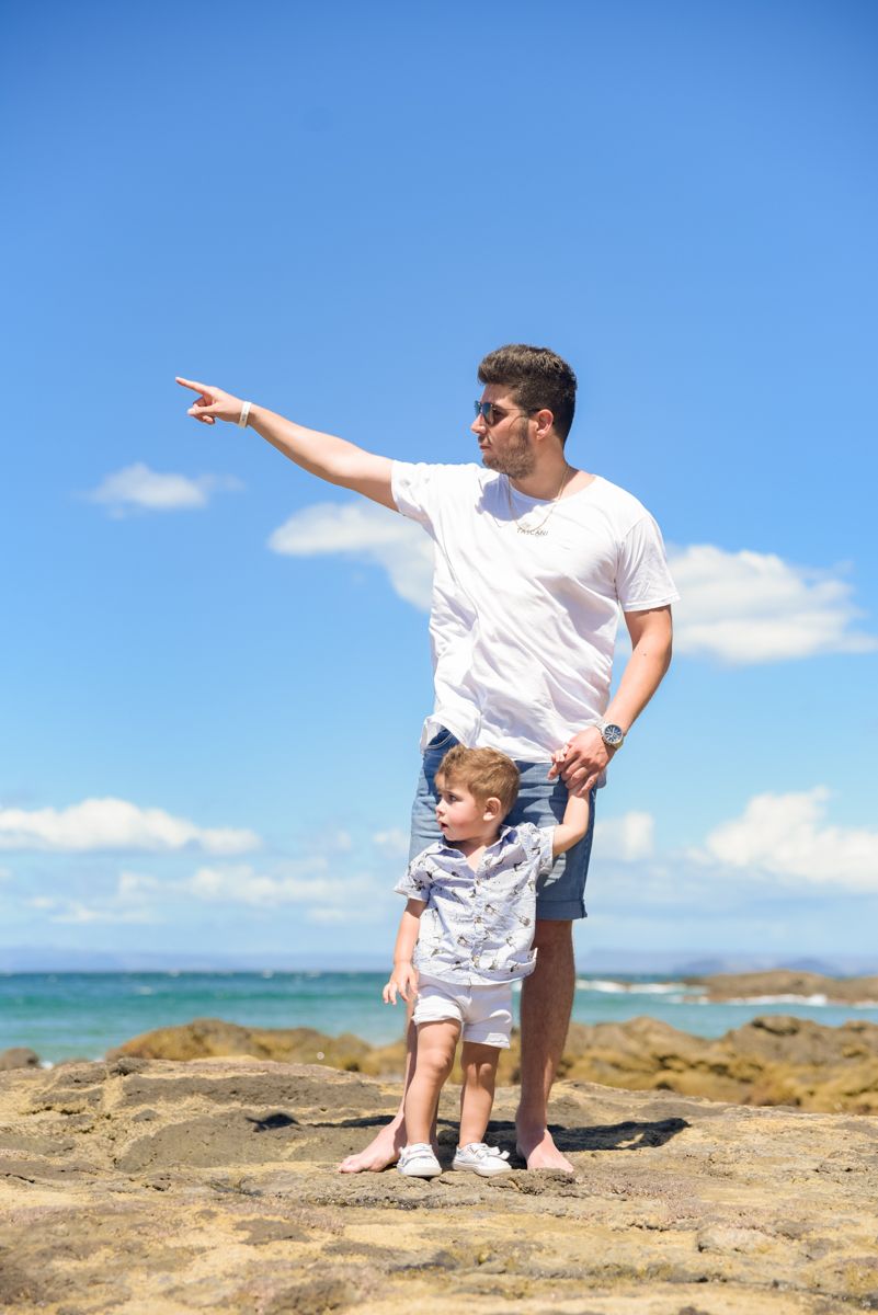Two people standing on rocky coastal outcrop pointing at ocean view on sunny day with blue sky and clouds.