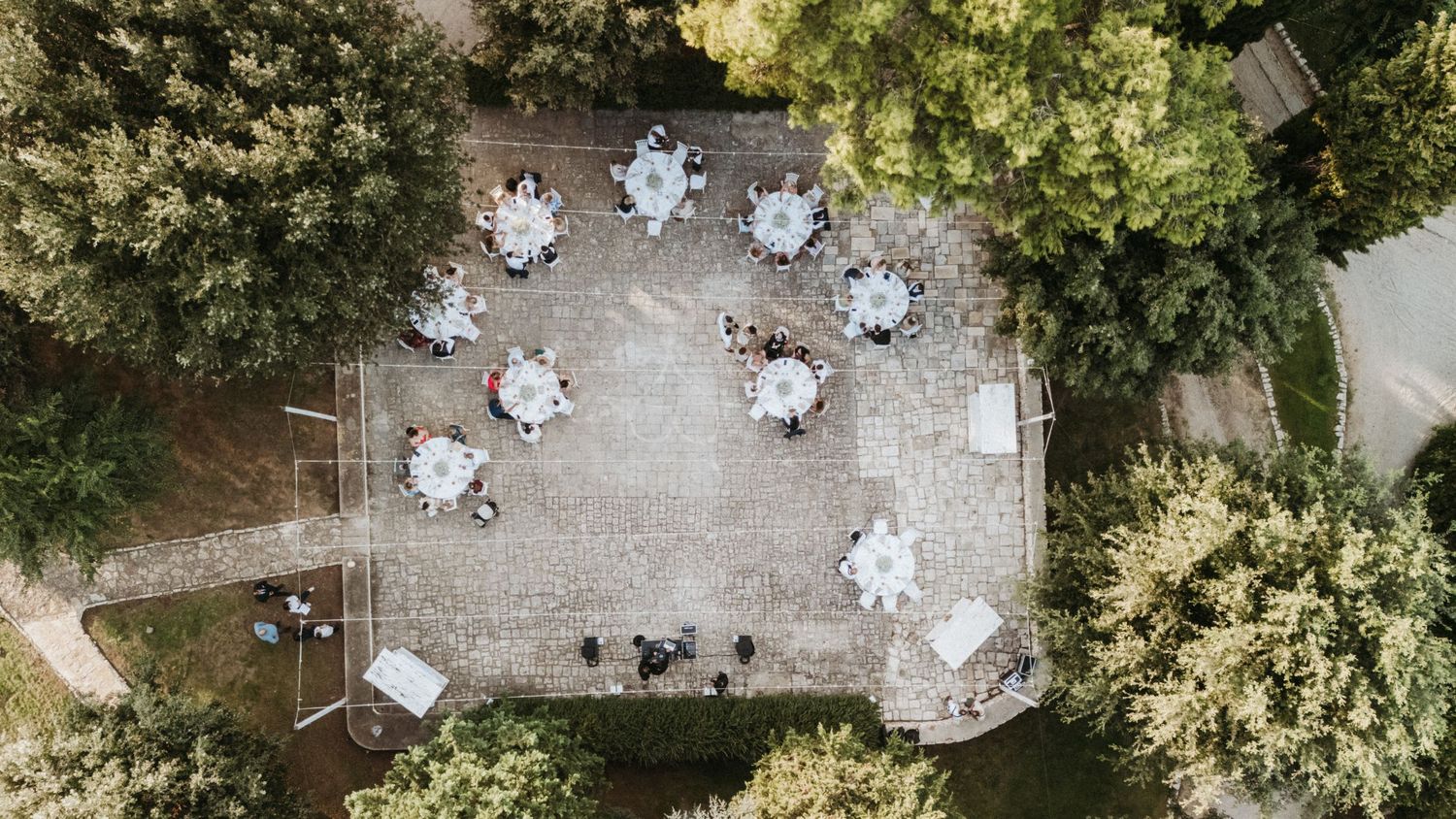 Aerial view of a wedding reception with white tables and chairs arranged under lush green trees in a garden setting.