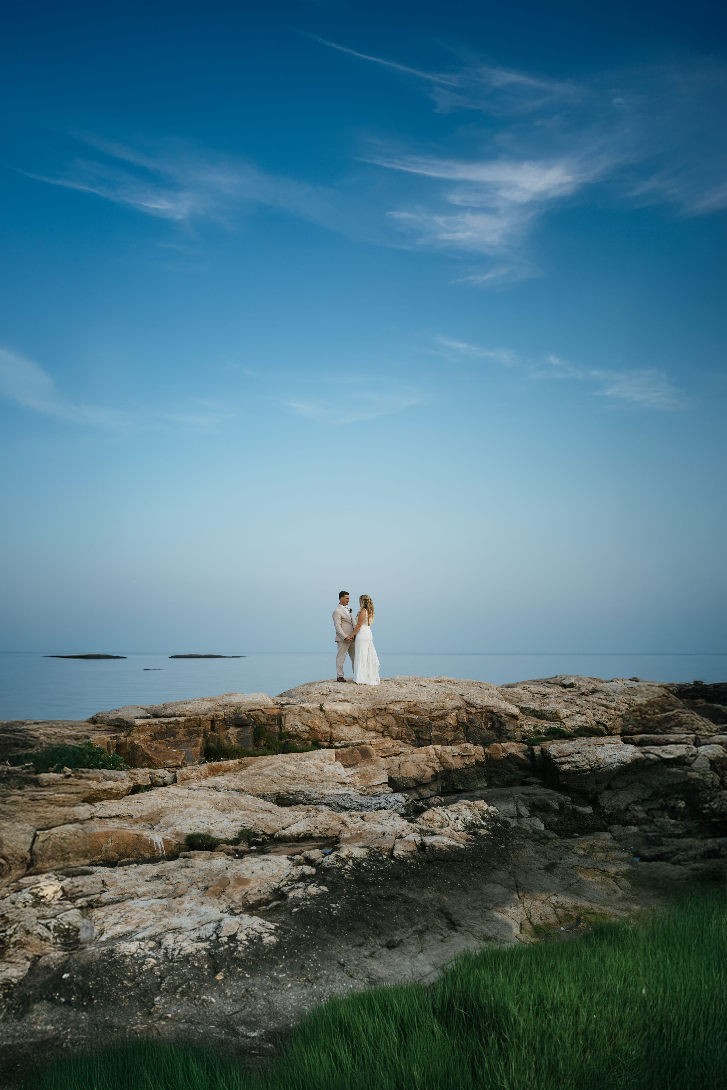 A wedding couple stands on coastal rocks against a bright blue sky and calm ocean waters.