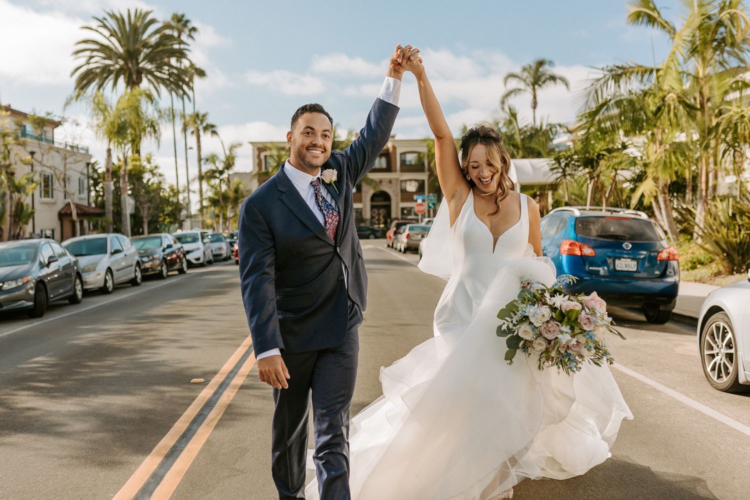 Joyful newlyweds celebrate on a sunny street, bride in white gown with bouquet, groom in navy suit, palm trees behind in La Jolla.