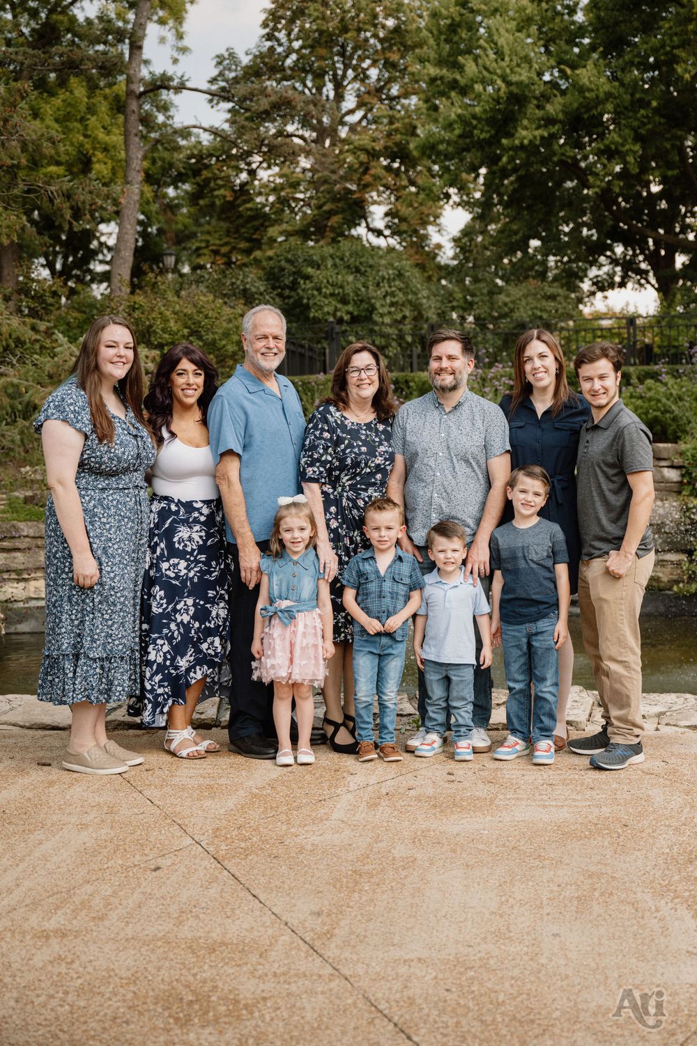 Large multigenerational family portrait outdoors near a pond, smiling together with lush green trees in the background.