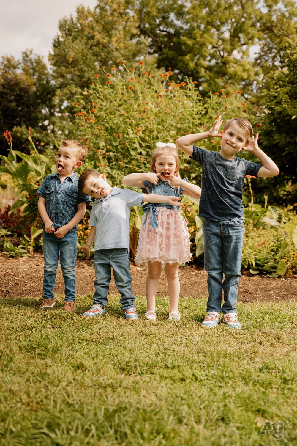 Four young children making silly faces and gestures while posing together on grass in front of colorful garden flowers.
