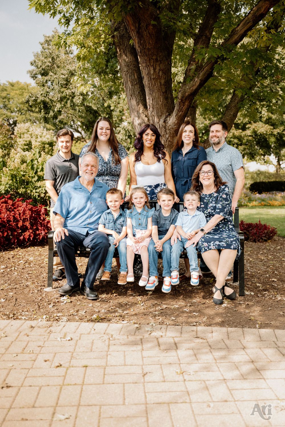 Large family portrait outdoors under a tree, with adults standing and children seated on a bench in a park.