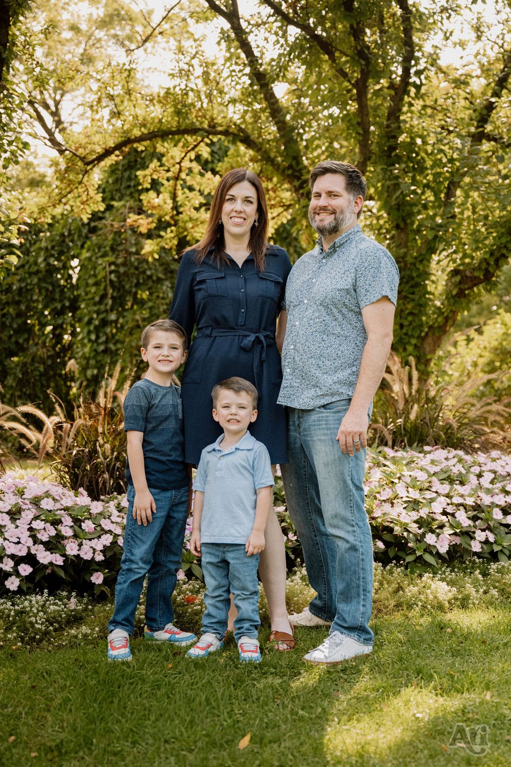 Family of four posing outdoors in a lush garden with pink flowers and green trees in the background.