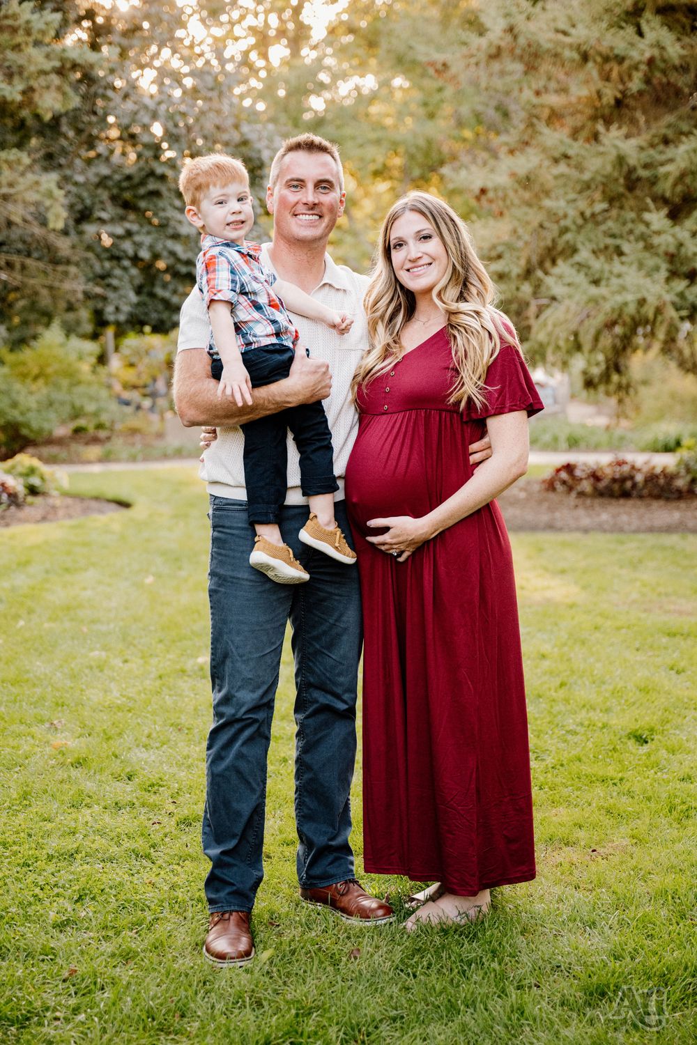 A smiling family poses outdoors, featuring a pregnant woman in a red dress, a man holding a toddler boy, on a green lawn.