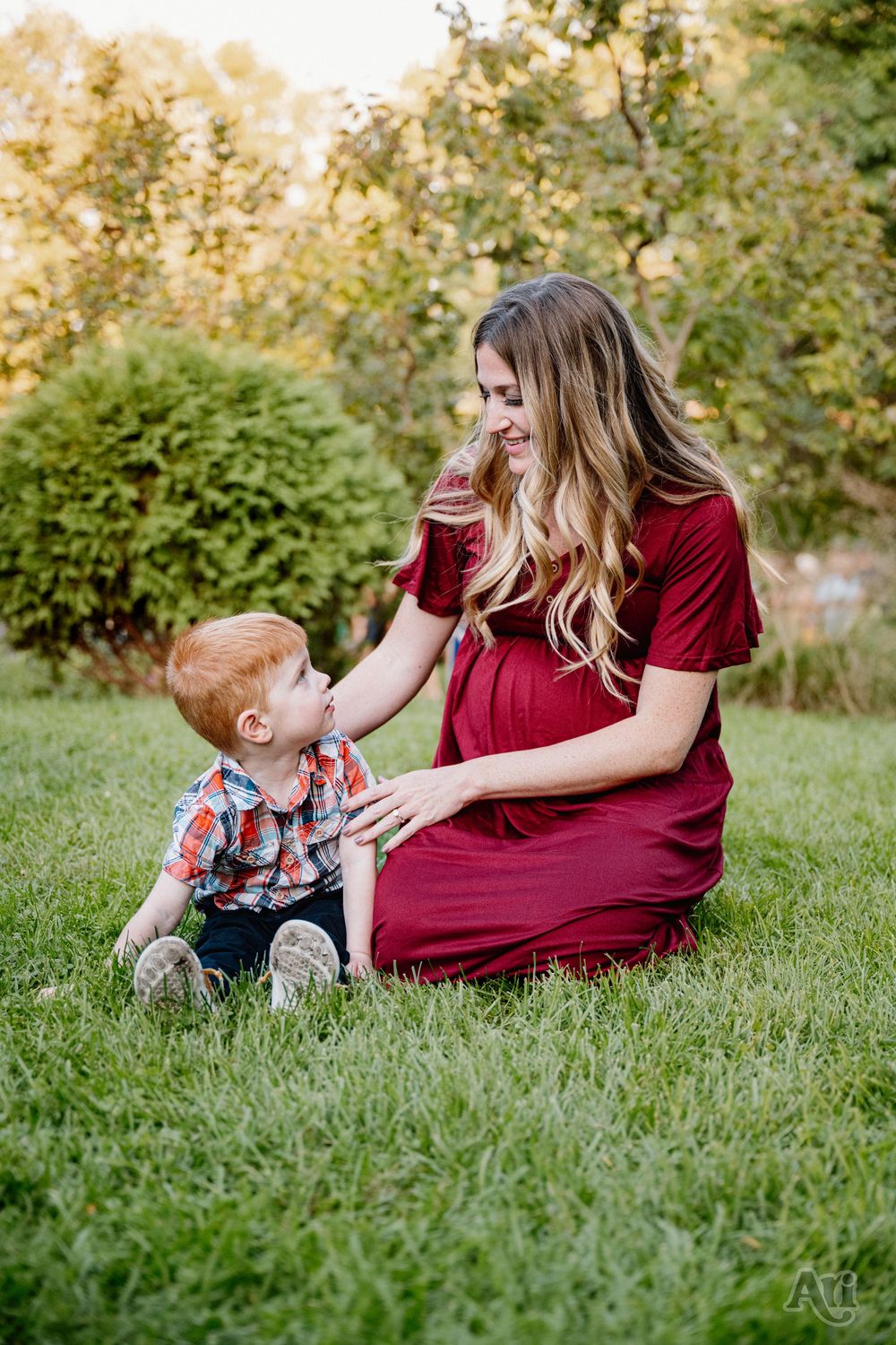 Pregnant woman in red dress sitting on grass smiling at red-haired toddler boy in a sunny green park.