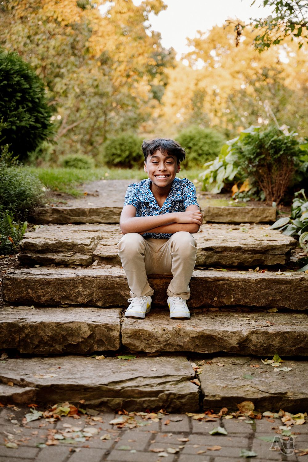 Smiling young boy sitting cross-legged on stone steps in a lush garden with autumn foliage.