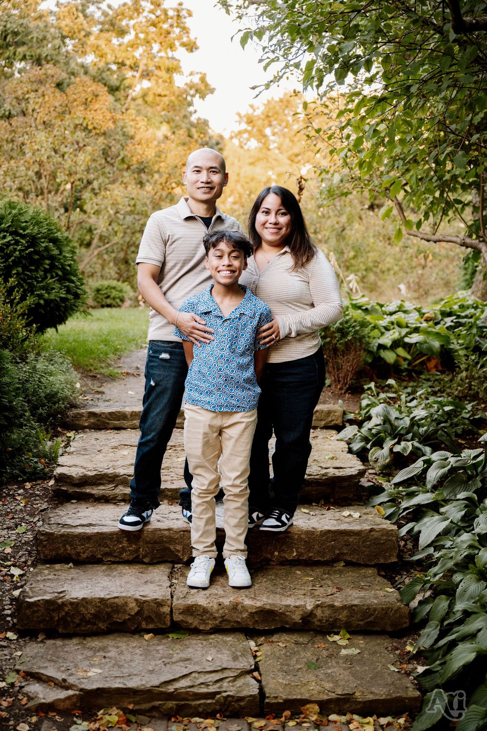 Family of three posing on stone steps surrounded by autumn foliage in a lush garden setting.
