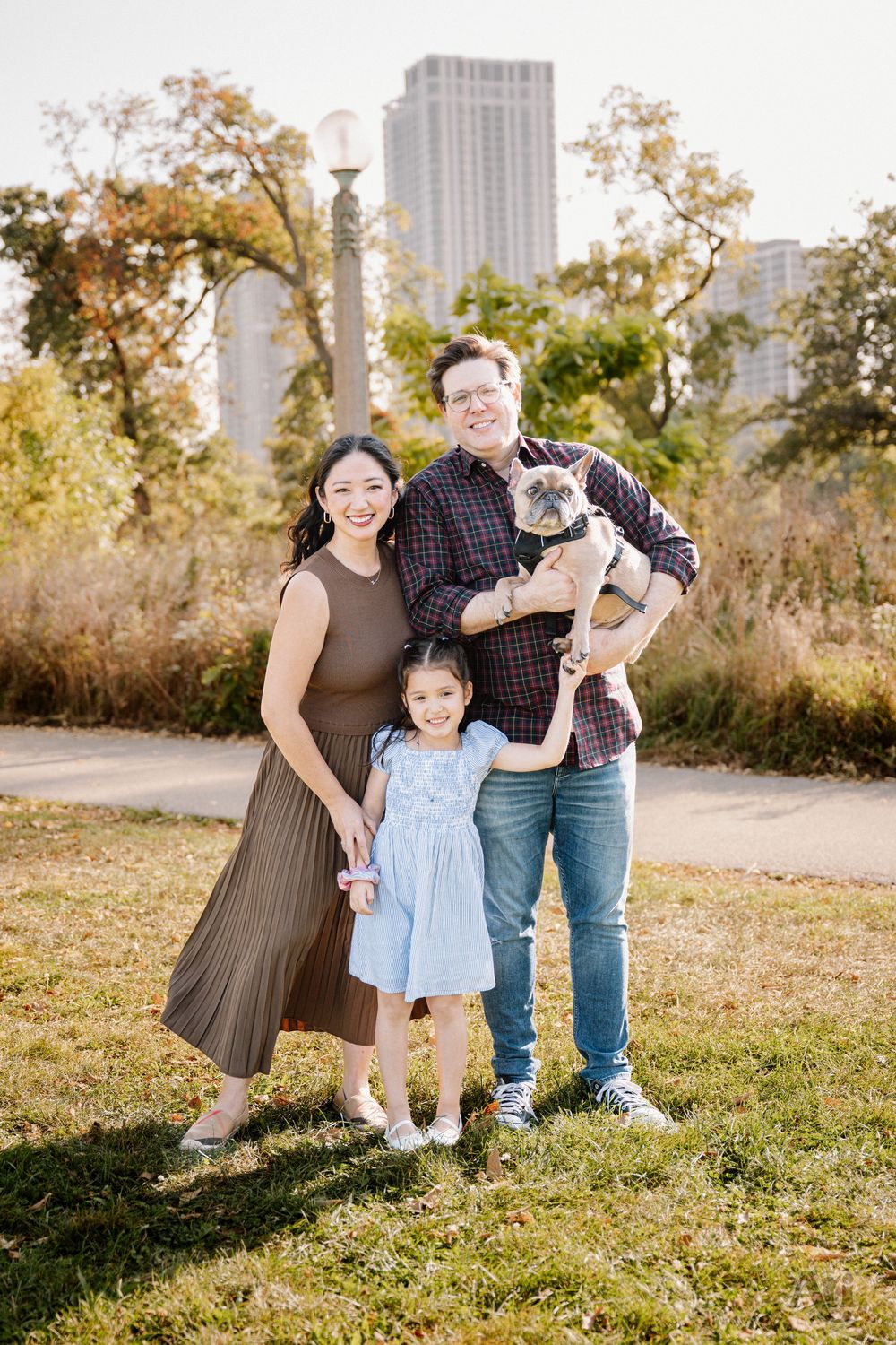 Family of three with a French Bulldog posing outdoors in a park with city skyline in the background.