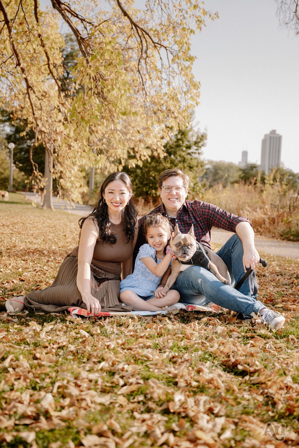 Family of three with a French Bulldog sitting on autumn leaves in a park, smiling warmly on a sunny fall day.