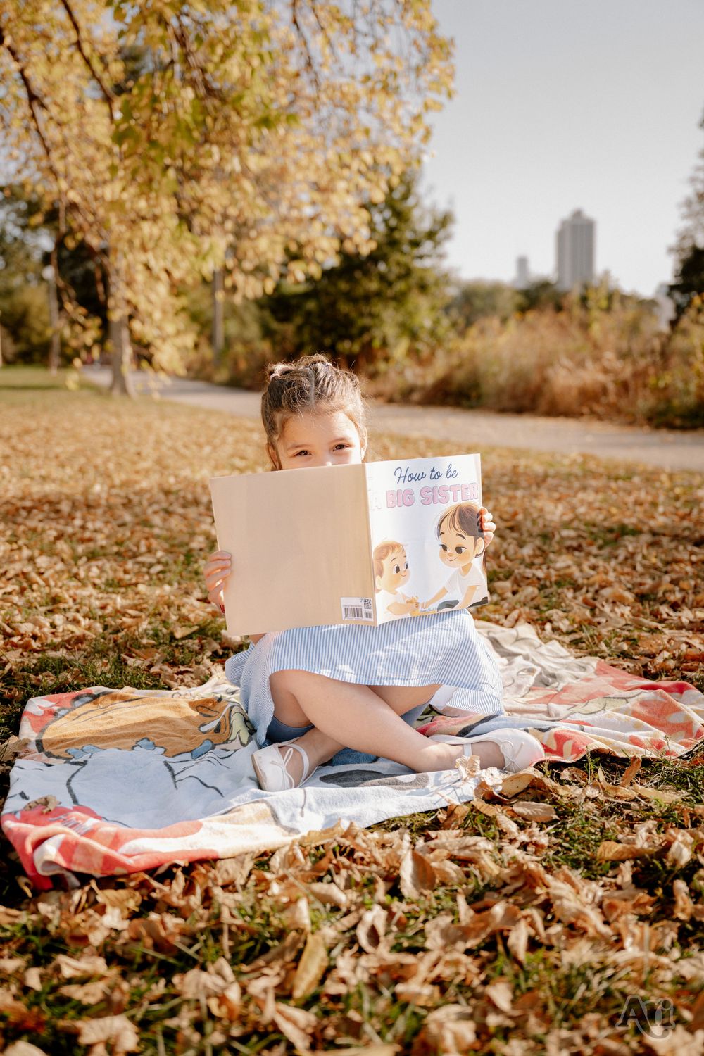 Young girl sitting cross-legged on a blanket in autumn park, reading a children's book outdoors.