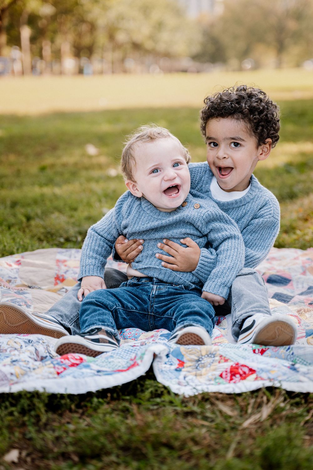 Two young boys in matching blue sweaters laughing on a colorful quilt in a park.