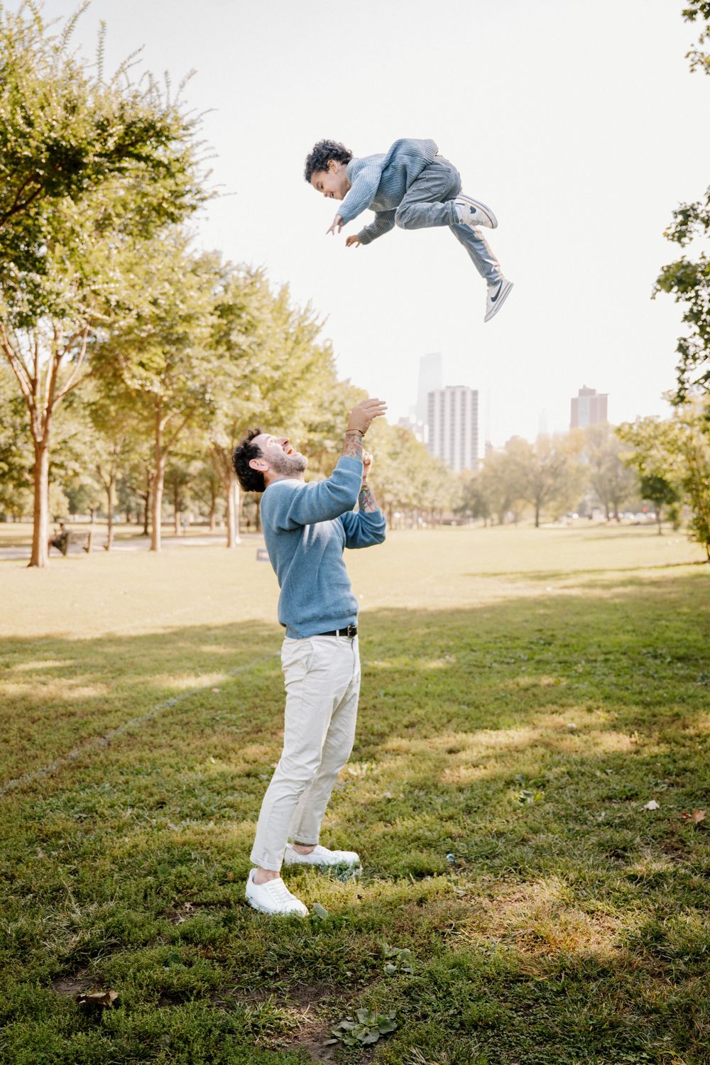 Father joyfully tosses young child into the air in a sunny green park, with city skyline visible in the background.
