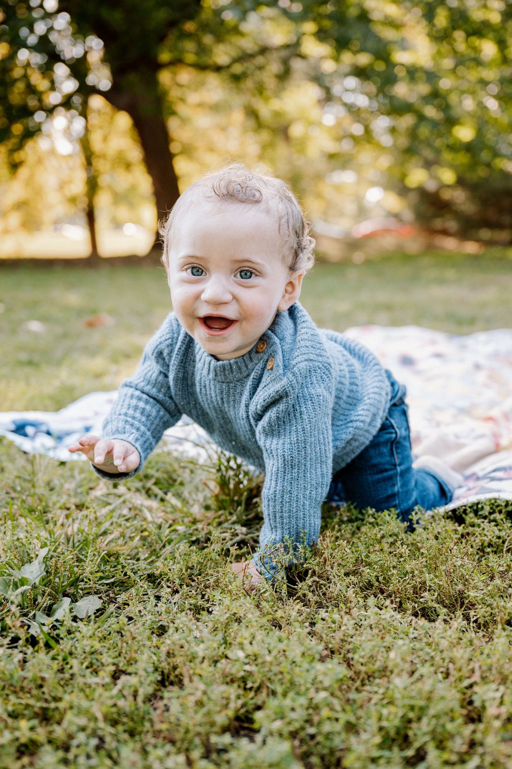 A happy baby in a blue knit sweater crawls on green grass in a sunny park setting.