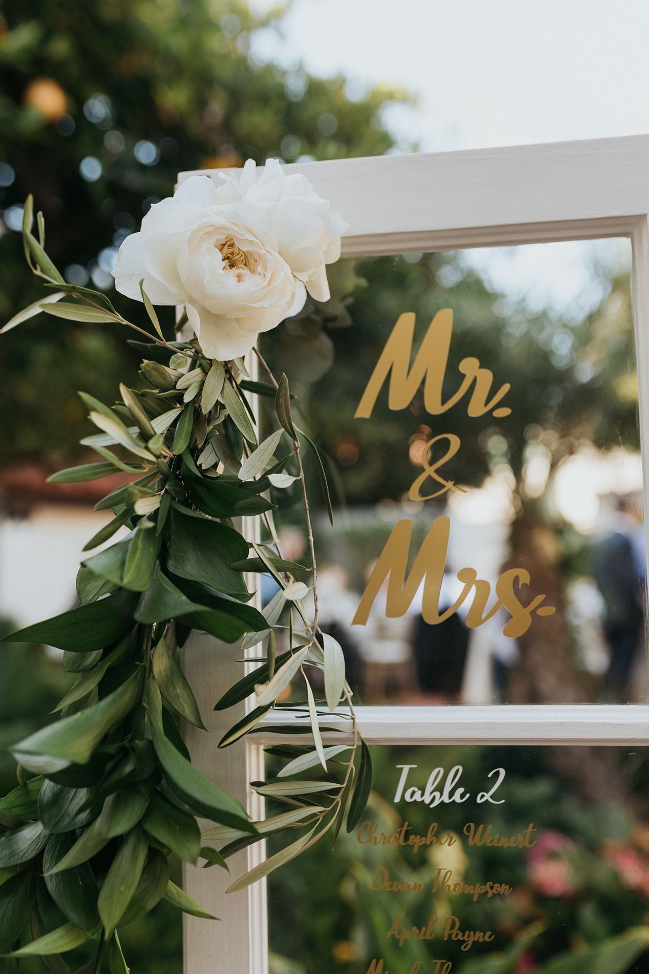 Elegant wedding seating chart on glass with gold Mr. & Mrs. lettering, white roses, and greenery garland decoration.