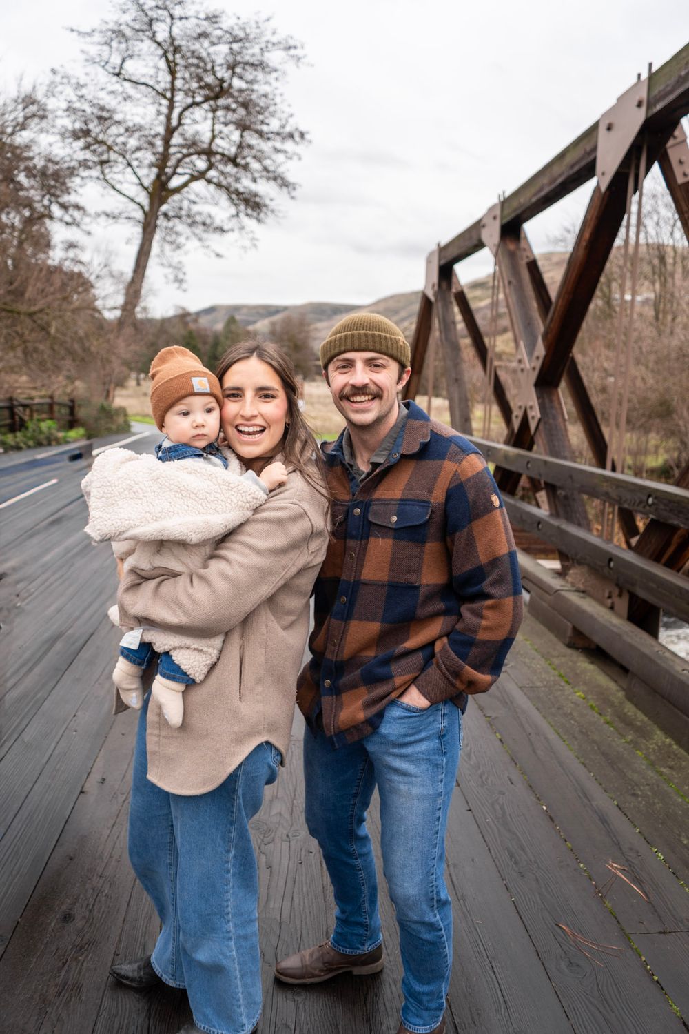 Young family with baby posing on a wooden bridge outdoors in winter, wearing casual flannel and beanies.