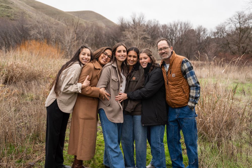 A happy family of six poses together smiling in a scenic outdoor field with rolling hills in the background.