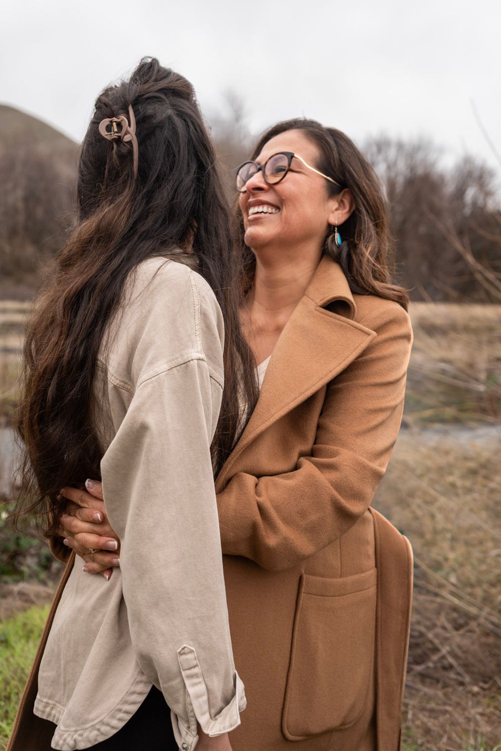 Two women in autumn coats embrace outdoors, smiling warmly in a natural setting on a cloudy day.