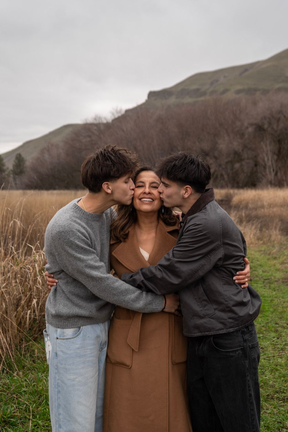 A woman smiles joyfully as two young men kiss her cheeks in a scenic outdoor field with hills behind.