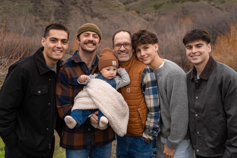 Six men of varying ages pose outdoors for a family portrait, with one holding a baby in a brown sherpa outfit.