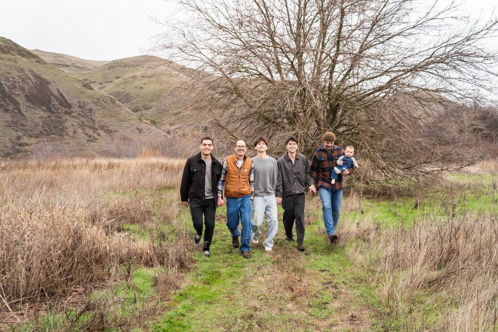 Group of young men hiking on a green trail path with rolling hills and bare trees in background.