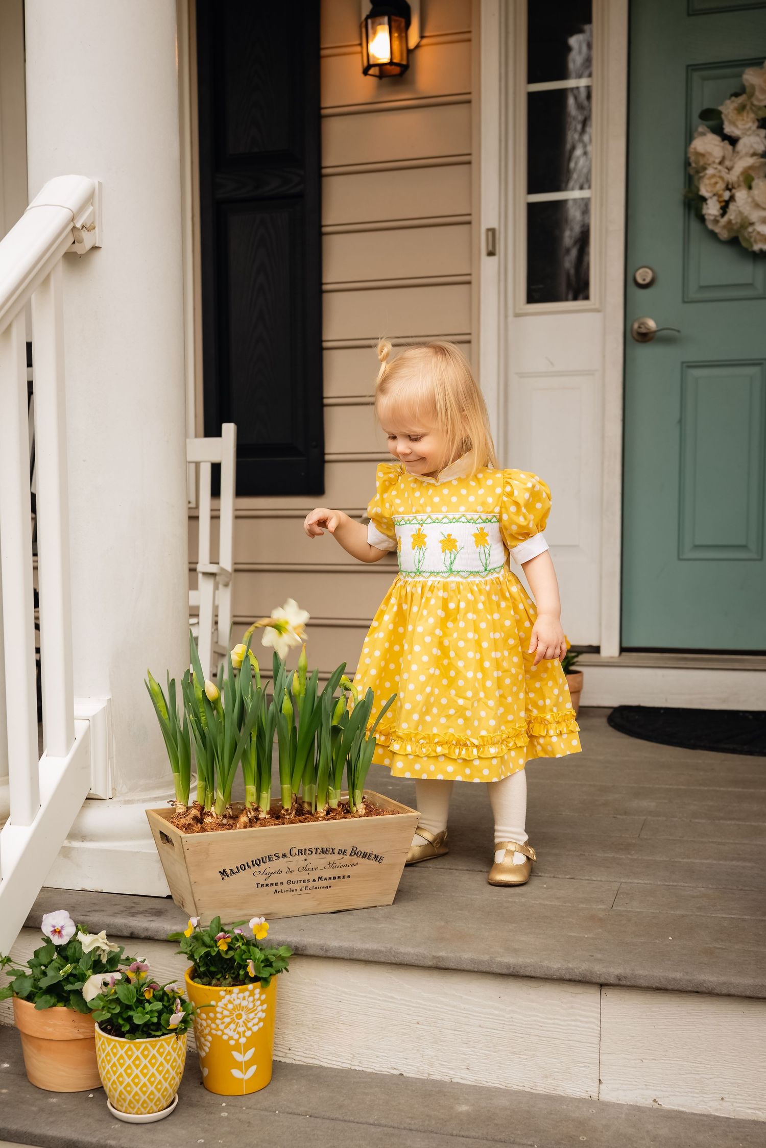 Toddler girl dressed in yellow daffodil dress reaches toward a crate of daffodils, captured by charlottesville family and baby photographer.