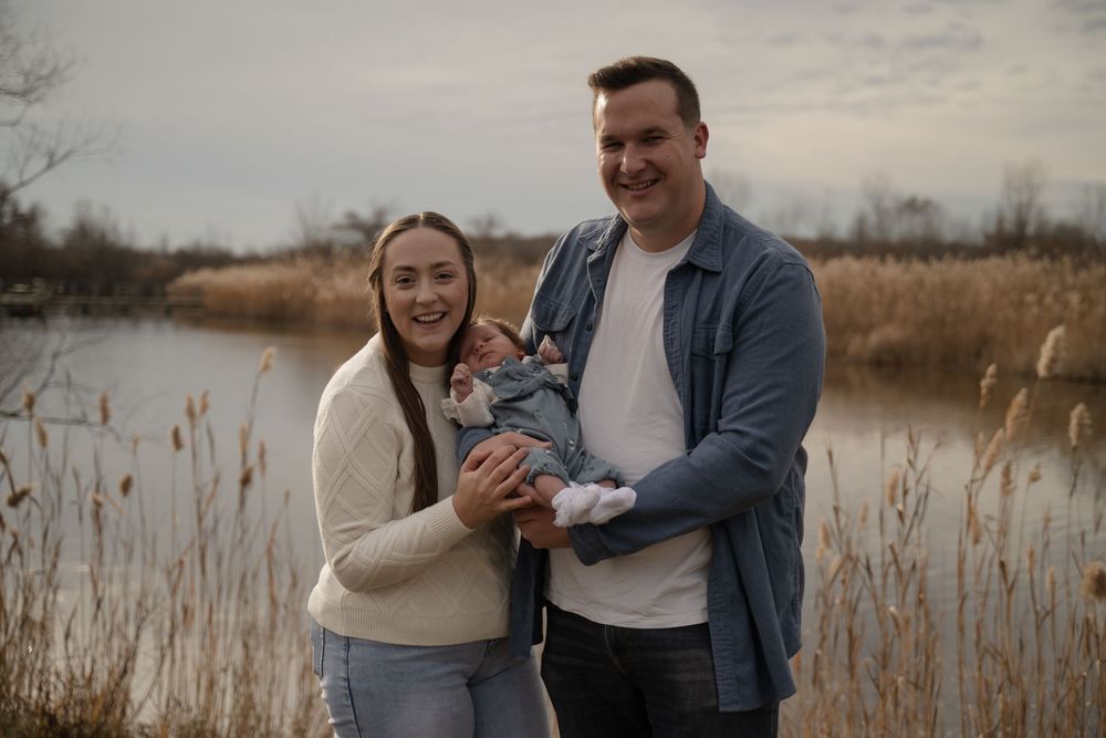 Happy young family with newborn baby posing near a lake surrounded by tall reeds.