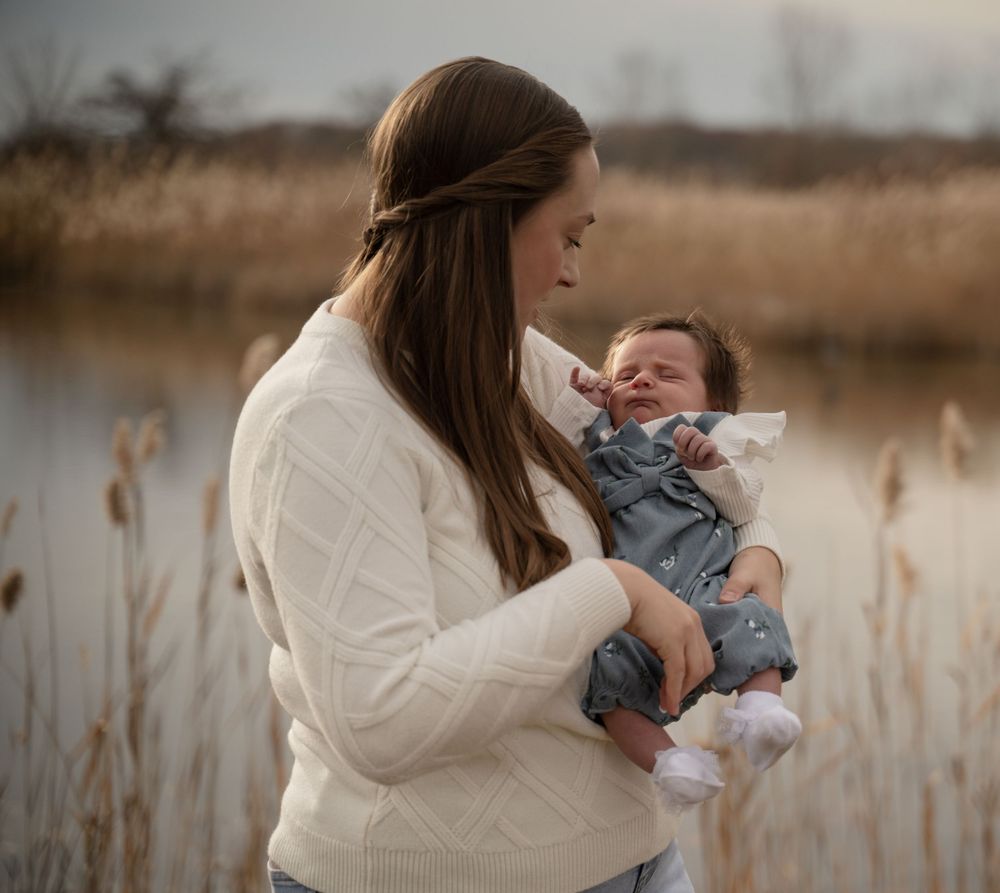 Mother in white sweater tenderly holds her newborn baby outdoors near a serene lake.