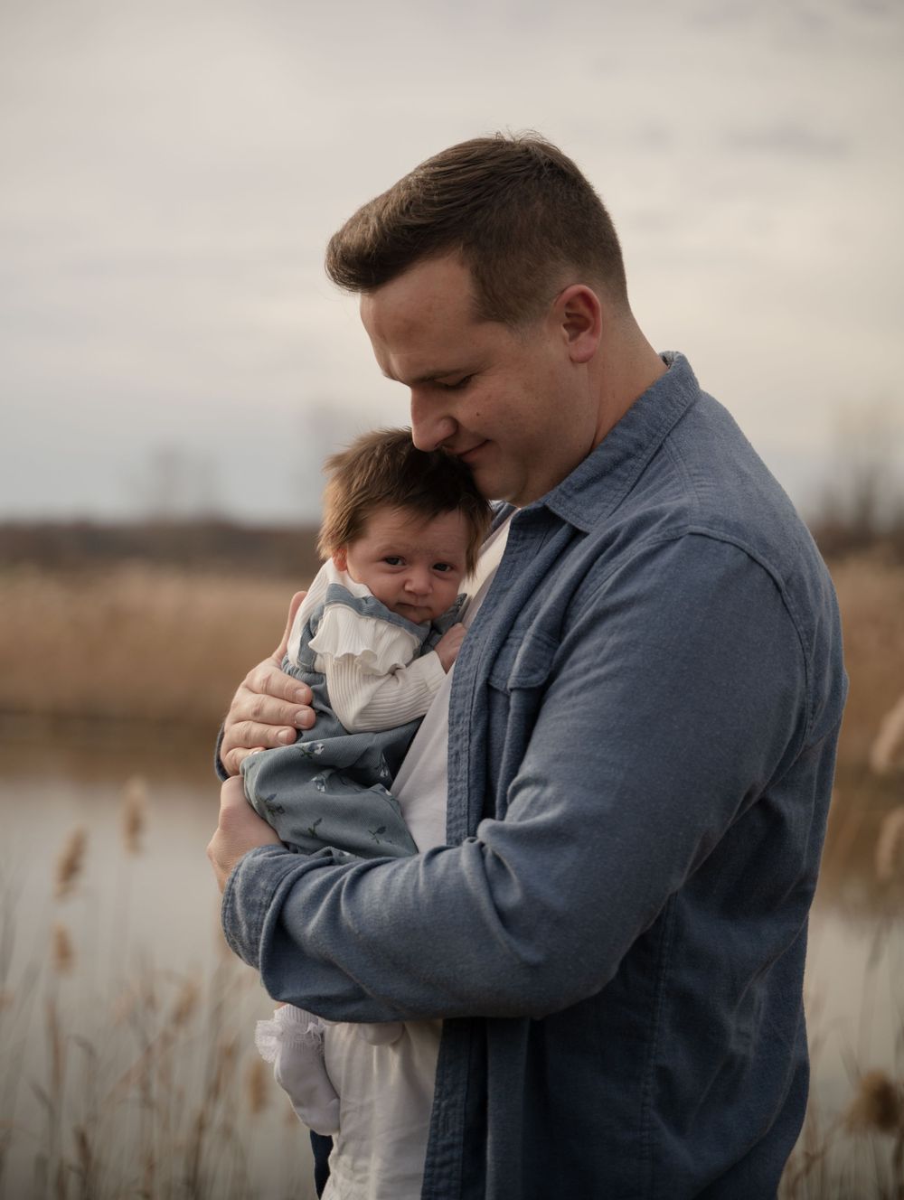 A father tenderly holds his newborn baby outdoors near a serene lake with tall reeds in the background.